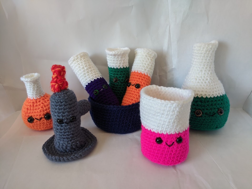 A set of crocheted toys. There are 3 test tubes, a beaker, 2 flasks and a bunsen burner in various colors.